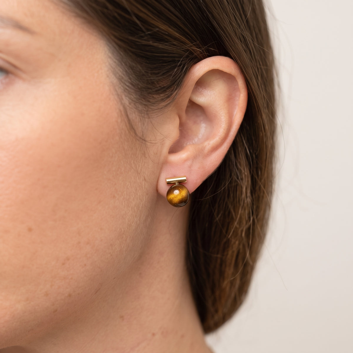 A close-up side profile of a woman's ear wearing a small gold post earring with a round amber-brown bead drop.