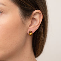 A close-up side profile of a woman's ear wearing a small gold post earring with a round amber-brown bead drop.