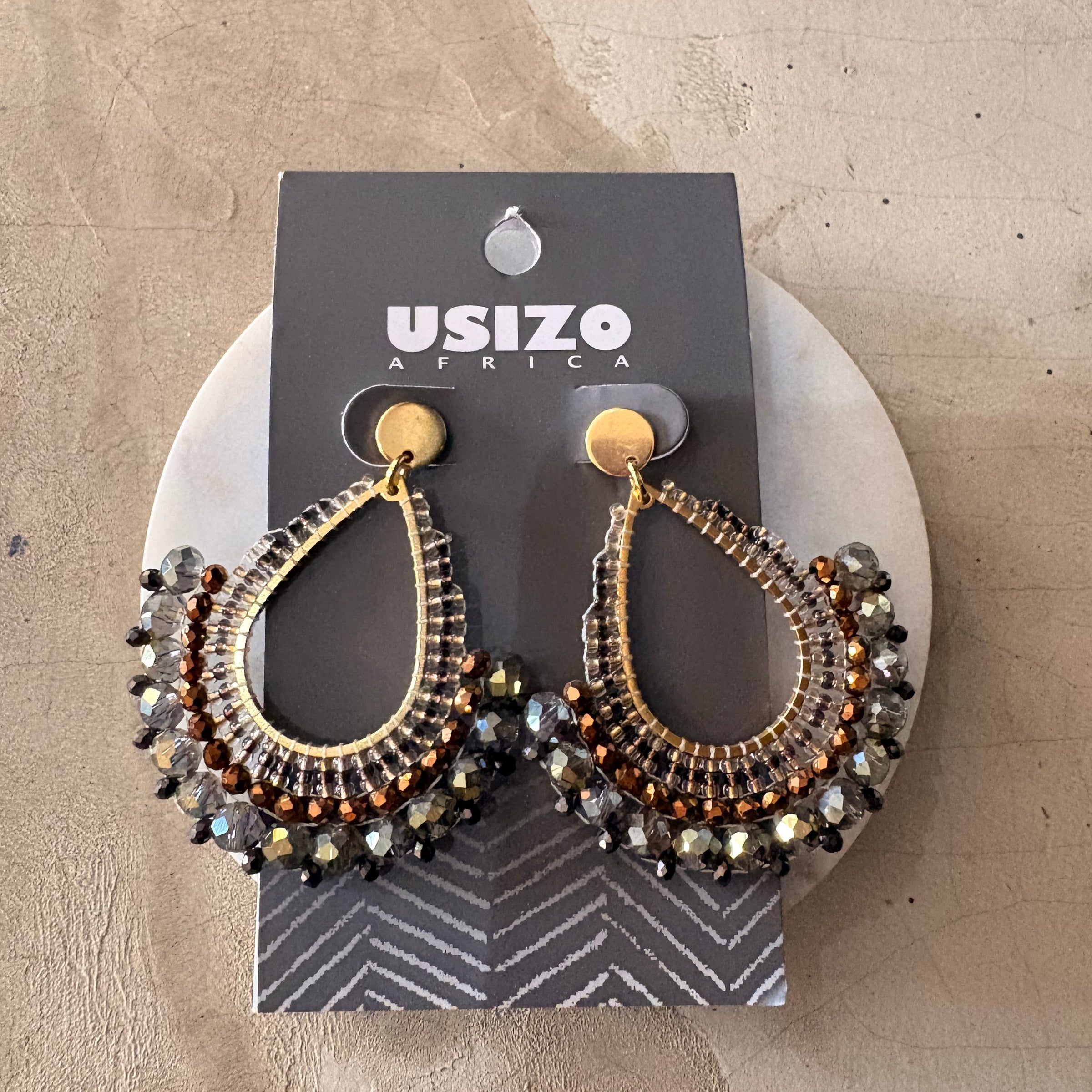 Pair of ornate earrings on a branded card against a textured beige background