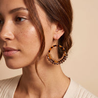 A close-up of a woman wearing a large hoop earring with a tortoiseshell pattern and a fringe of brown beads, set against a warm beige background.