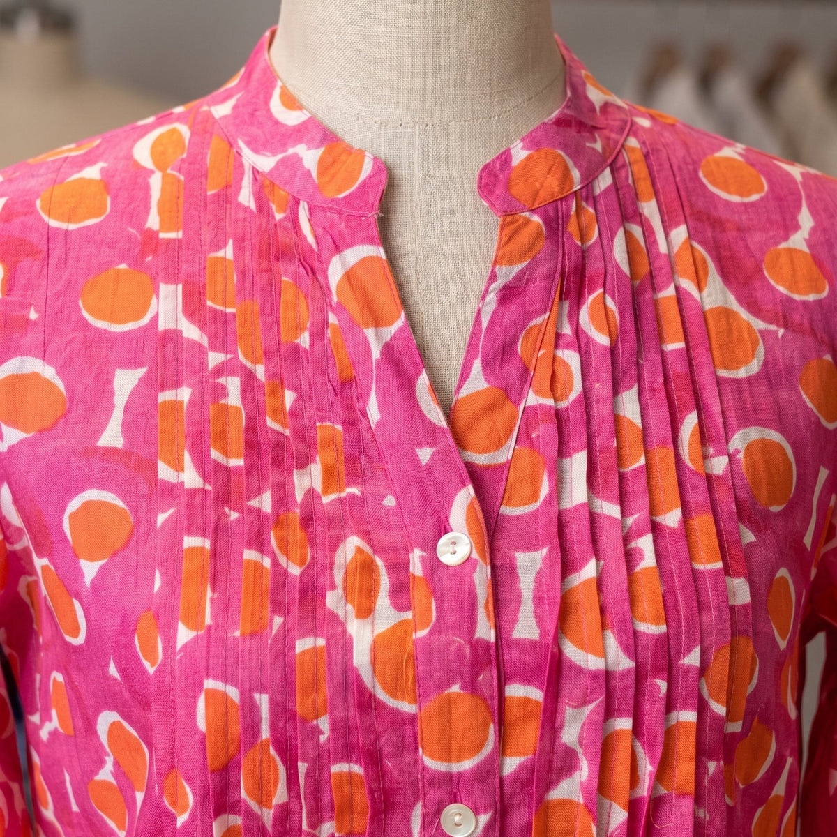 A close-up of a pleated pink blouse on a mannequin, featuring an orange and white abstract pattern and a split neckline.
