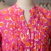 A close-up of a pleated pink blouse on a mannequin, featuring an orange and white abstract pattern and a split neckline.