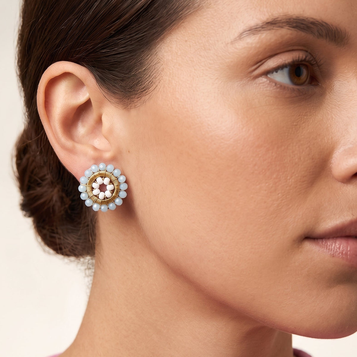 A close-up of a woman wearing a round gold and white beaded stud earring, shown from the side against a soft beige background.