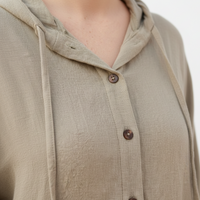 Close-up of a person wearing a light khaki hooded button-front top with drawstrings and brown buttons, photographed outdoors with a softly blurred background.